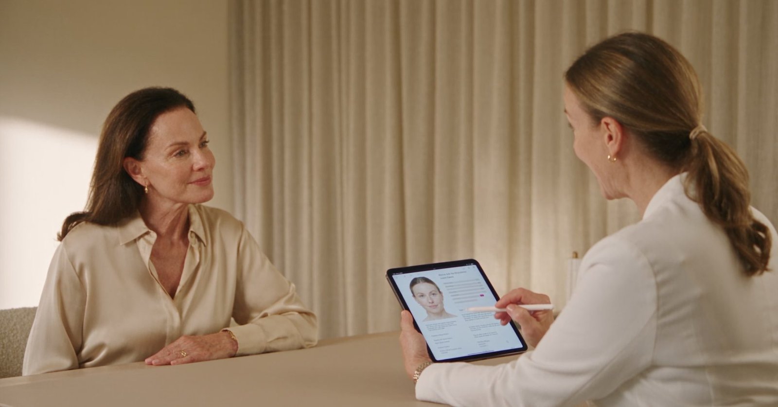 A clinician sits across a consulting table from a client in a calm treatment room, showing her a digital consultation record and treatment plan on a tablet while holding a stylus.